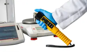 Ręka w niebieskiej rękawiczce trzyma żółty alkomat. W tle waga laboratoryjna.