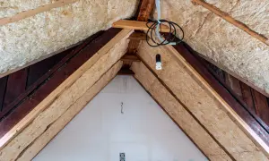 Dämmmaterial wird in einem Dachboden verlegt, um das Haus zu isolieren. Holzbalken und Kabel sind sichtbar.