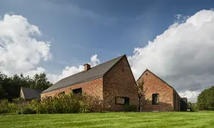 Nowoczesny blok z cegły z ciemnym dachem, otoczony zielonym trawnikiem i bujną roślinnością.
