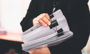 Osoba w czerni trzyma stos dokumentów, wśród których znajduje się ewidencja przebiegu pojazdu dla celów VAT.