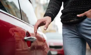 Mano tocando la manija de un vehículo rojo, con un suéter y jeans. Parece que el conductor está a punto de entrar.