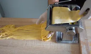 Ręce w rękawiczkach formują prosta pasta fresca na maszynce do makaronu. Świeży, żółty makaron opada na drewniany blat.