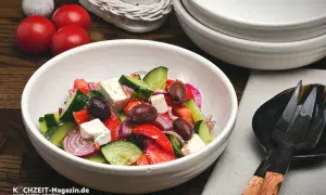 Frischer griechischer Salat mit Tomaten, Gurken, roten Zwiebeln, Oliven und Feta in einer weißen Schüssel.