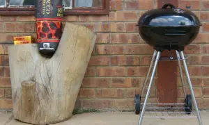 Grill stoi obok pnia, a torba z węglem drzewnym wisi na pniu. Odległość grilla od granicy działki jest bezpieczna.