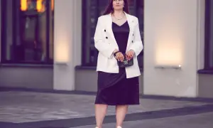 Eine Frau in einem schwarzen Kleid und weißem Blazer, die zeigt, was man zum Musical anzieht. Sie trägt weiße Sneaker und eine kleine Handtasche.