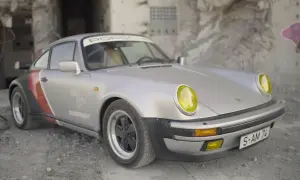 Srebrny Porsche 911 z żółtymi reflektorami, w stylu cyberpunk, stoi na tle zniszczonej ściany z graffiti.