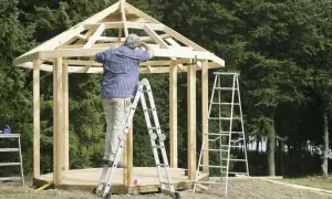 Man bygger lusthus i trä. Takstolar monteras på en stege.