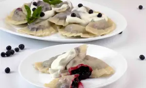 Pyszne pierogi z jagodami, polane śmietaną i ozdobione miętą. Na talerzu widać też kilka świeżych jagód.