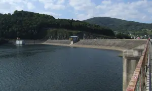 Betonowa zapora tworzy rozległe, spokojne tresna jezioro. W oddali widać zalesione wzgórza i niewielką wieś.