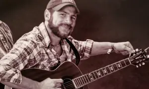 Kyle Shearer, a bearded man in a plaid shirt and cap, strums his acoustic guitar, ready to play a tune.
