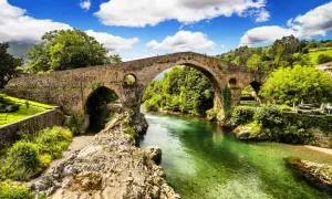 Majestuoso puente romano asturias sobre un río de aguas cristalinas, rodeado de exuberante vegetación y un cielo azul con nubes blancas.