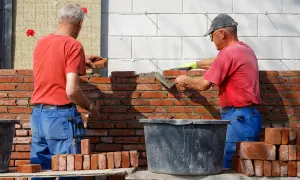 Dwóch budowlańców stosuje zasady murowania z cegły pełnej, układając cegły na zaprawie.