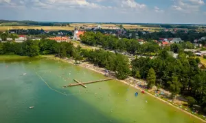 Tłum ludzi wypoczywa nad jeziorem wielewskim. Woda jest zielona, a na brzegu widać plażę i las. W oddali majaczy kościół.