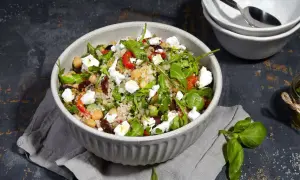 Bunter Quinoa Salat mit Kichererbsen, Feta, Oliven und Rucola, serviert in einer Schüssel. Daneben ein Glas mit Kräuter Dressing.
