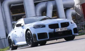 Jasnoniebieskie BMW M2 z czarnymi felgami i spoilerami, na tle przemysłowych rur. Sprawdź aktualną bmw m2 cena.