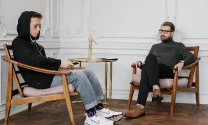 Chłopak w bluzie z kapturem i słuchawkami, trzymający pilota, siedzi naprzeciwko mężczyzny w okularach. Rozmowa o spadku po rodzicach.