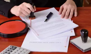 Dłoń z pieczęcią nad dokumentem, na którym widnieje napis "LAST WILL AND TESTAMENT". Obok poduszka z tuszem i długopis. To akt notarialny o poddaniu się egzekucji.