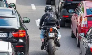 Motocyklista w kasku i plecaku przeciska się między samochodami w korku. Widać znaki motocyklistów na tablicy rejestracyjnej.