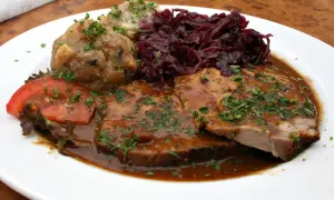Pyszne jedzenie w Niemczech: pieczeń wieprzowa w sosie, kluski ziemniaczane, czerwona kapusta i pomidor.