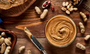 Cremige Erdnussbutter in einer Schüssel, daneben Erdnüsse und ein Messer. Erdnussbutter gesund und lecker!