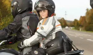Uśmiechnięte dziecko na motocyklu, siedzące za kierowcą, w kombinezonie i kasku, gotowe na przygodę.