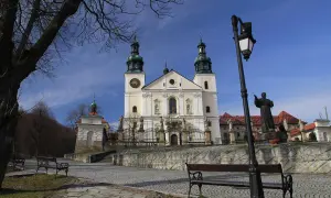 Kalwaria Zebrzydowska: barokowy kościół z wieżami, plac z ławkami i pomnik.