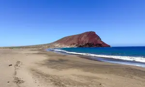 Plaże Teneryfy z charakterystyczną czerwoną górą w tle. Spokojne fale obmywają piaszczysty brzeg.