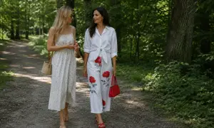 Dwie kobiety spacerują po leśnej ścieżce. Jedna w sukience w drobne kwiatki, druga w eleganckich spodniach w kwiaty z dużymi czerwonymi różami.