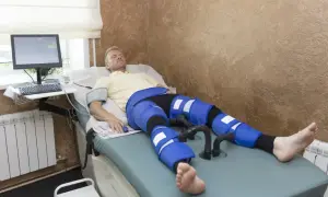 Mężczyzna leży na łóżku rehabilitacyjnym, poddając się zabiegowi limfodrenaż. Na nogach ma założone specjalne mankiety.