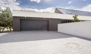 Nowoczesny dom z ciemnym garażem, którego brama otwiera się ale nie zamyka. Podjazd z kostki brukowej, biały murek i drzewa.