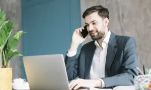 Uśmiechnięty pkd agent ubezpieczeniowy rozmawia przez telefon, pracując przy laptopie.