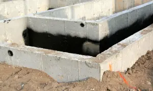 Surowe ściany fundamentowe z betonu, częściowo pokryte czarną izolacją przeciwwilgociową, otoczone ziemią.