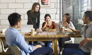 Team discute progetti, con una donna che indica documenti e un uomo che parla. Si discute di trasferte in busta paga.