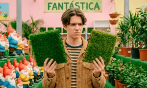 Joven indeciso compara dos muestras de césped artificial, buscando el mejor césped artificial del mercado. Detrás, gnomos de jardín y plantas.