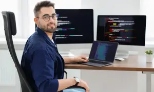 Typowy programista w okularach siedzi przy biurku z trzema monitorami wyświetlającymi kod.