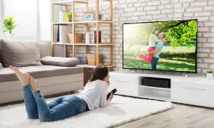 Une jeune femme se repose sur un tapis blanc, télécommande en main, pour regarder la tv sans connexion internet. L'écran affiche un couple s'enlaçant.