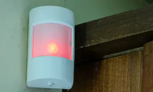 Biały czujnik ruchu z czerwoną diodą, zamontowany na ścianie obok drewnianej framugi. Pokazuje, jak działa czujnik ruchu.