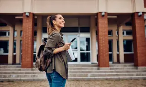 Uśmiechnięta studentka z plecakiem i telefonem przed budynkiem uczelni. Zastanawia się, jakie studia po rozszerzonym angielskim wybrać.