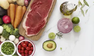 Un corte de carne magra, rodeado de verduras frescas como zanahorias, brócoli, tomates y aguacate, junto a aceite de oliva y limón.