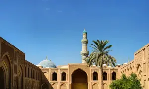 Ein Minarett und eine Kuppel einer Moschee ragen über traditionelle Gebäude mit Bögen und Palmen. Einblick in die reiche irakische Kultur.