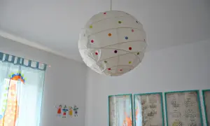 Jak odnowić papierową lampę? Biała lampa wisząca w kropki, obok niebieskich zasłon z dinozaurem i rysunków dzieci.