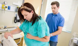 En gravid kvinna håller sig om magen, kanske upplever hon gbs gravid sammandragningar. Hennes partner stöttar henne i sjukhusmiljö.