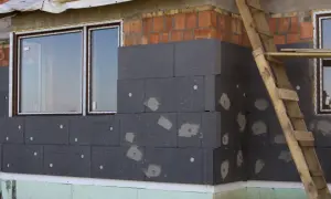 Budowa domu w toku, trwa ocieplanie ścian zewnętrznych styropianem. Widać okna i fragment rusztowania.