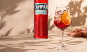 Orzeźwiający Campari Spritz z plasterkiem pomarańczy i kostkami lodu, obok butelki Campari. Idealny na letnie popołudnie.