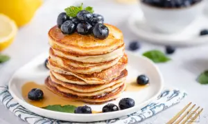 Puszyste placki z ricotty, polane syropem i ozdobione borówkami, wyglądają apetycznie. Obok leżą cytryny.