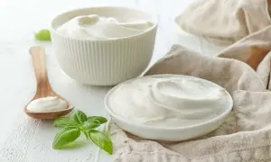 Gładki, kremowy jogurt naturalny w miseczkach i na drewnianej łyżce, ozdobiony listkami bazylii.