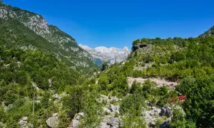 Grüne Hänge und felsige Gipfel in den albanischen Alpen unter strahlend blauem Himmel. Ein kleiner roter Steg führt über einen steinigen Bachlauf.