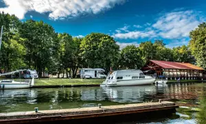 Malownicze pojezierza w Polsce: kampery i łodzie cumują przy brzegu, w tle drewniana restauracja i bujna zieleń.