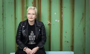 Ola Liljedahl, med kort blont hår och glasögon, sitter framför en sliten grön vägg. Hon bär en svart läderjacka och en t-shirt med texten "THE GREAT NORRLAND NATUREN".