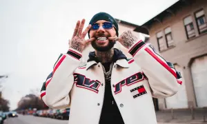 Malik Montana z uśmiechem i błyszczącymi zębami, w kurtce Tommy Jeans. Jego wzrost jest imponujący, a tatuaże dodają mu charakteru.